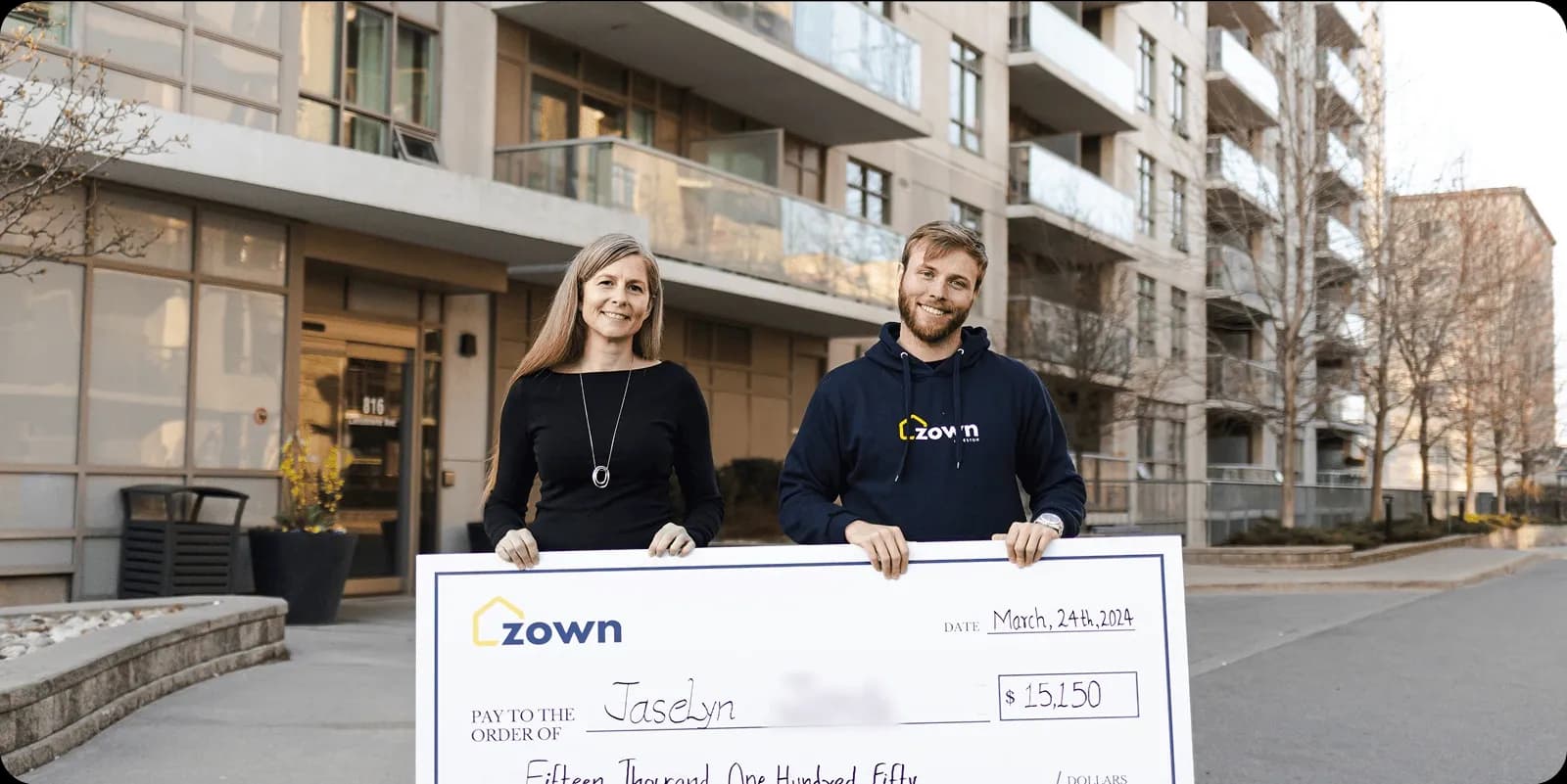 Homeowners standing in front of a condo building holding a large Zown cheque for down payment assistance, representing real clients who received financial support and successfully purchased their home.